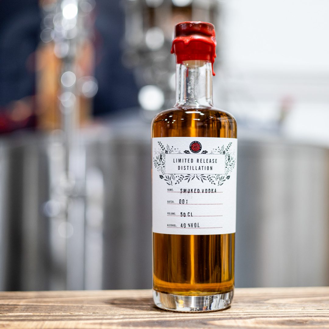 Spirit of Wales - Distillery Release - Smoked Welsh Vodka 3 Spirit of Wales Spiced Rum on a wooden bar with a copper still with a stainless steel still in the background.